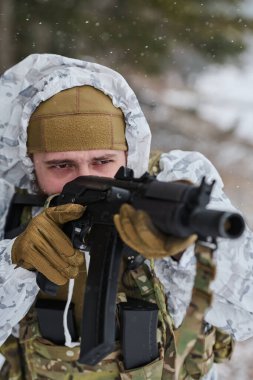 Beyaz kış kamuflajlı odaklanmış bir asker, soğuk hava askeri eğitimi veya devriye sırasında taktik yelek, eldiven ve bere giyerken kar altında bir saldırı tüfeğini hedefler..