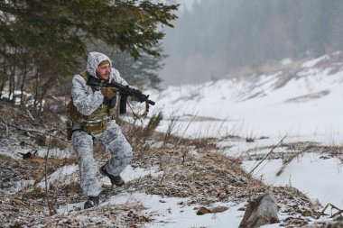 Beyaz kışlık kamuflaj ve taktik teçhizat kuşanmış silahlı bir asker, karlı bir orman sınırında dikkatli bir şekilde hareket ederek, engebeli ve ücra bir yerdeki soğuk hava askeri eğitim devriyesi sırasında tüfeğini doğrultuyor.