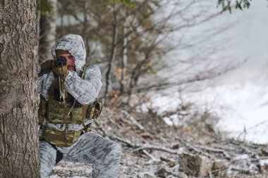 Kamuflajlı bir asker karlı bir ormandaki bir ağacın arkasında diz çökerek kış taktik eğitimi ve keşif sırasında tüfeği doğrultuyor. Sahne odaklanmayı, gizliliği, disiplini ve soğuk havayı aktarıyor.