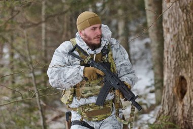 Beyaz kışlık kamuflaj ve taktik teçhizat giyen silahlı asker, karlı bir ormanda ihtiyatlı bir şekilde hareket ediyor, bir devriye ya da eğitim operasyonu sırasında elinde tüfekle, soğuk koşullarda odaklanmış ve tetikte..