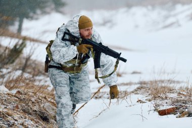 Beyaz kışlık kamuflaj ve taktik teçhizat takan bir asker karlı dağlık arazide devriye geziyor, elinde tüfekle serin bir havada sürüklenirken ve seyrek bir bitki örtüsü altında geziniyor.