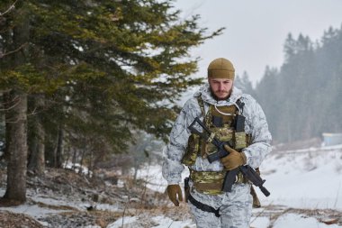 Kış kamuflajlı ve taktik teçhizatlı silahlı askerler hafif kar yağışı sırasında karlı bir ormanda yürürler, uzak bir yerdeki ihtiyatı, hayatta kalma eğitimini ve soğuk hava askeri operasyonlarını aktarırlar.