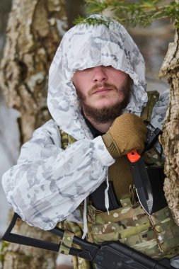 Beyaz kış kamuflajı giymiş sakallı bir asker, karlı bir ormanda devriye gezerken taktiksel bir savaş bıçağını kavrıyor..