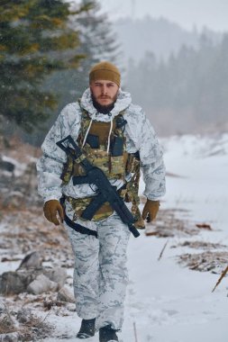 Beyaz kış kamuflajı ve taktik teçhizatı giyen bir asker karlı bir ormanda devriye geziyor, elinde tüfekle ve bulutlu havalarda soğuk hava teçhizatı ve hafif kar yağışı altında yürüyor..