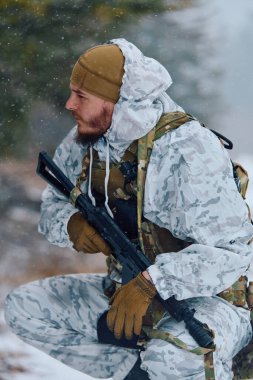 Beyaz kış kamuflajlı ve taktik teçhizatlı bir asker kar yağarken çömelir, soğuk hava devriyesi sırasında karlı bir ormanı incelerken tüfeği kavrar ya da tatbikat yapar..