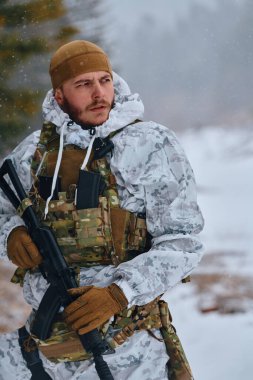 Beyaz kış kamuflajı ve taktik yelek giyen bir asker, karlı bir araziyi tararken bir tüfeği kavrarken, tetikte, hazır ve soğuk hava askeri operasyonlarını ormanlarda sürdürüyor.