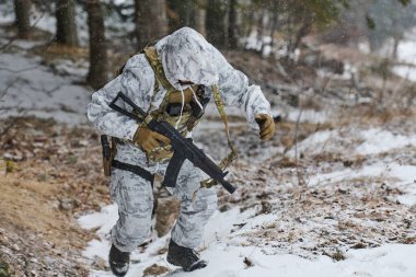 Beyaz kış kamuflajı giyen bir asker, bir tüfek ve taktik teçhizat taşıyan karlı bir orman yamacına tırmanıyor, ormanlık alanda soğuk hava eğitimi veya devriye sırasında ihtiyatlı hareket ediyor..