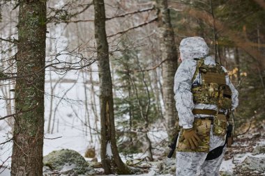 Beyaz kış kamuflajı giyen bir asker, karlı bir ormanda devriye geziyor, hafif kar yağışı sırasında ormanlık alanda keşif yaparken taktik bir sırt çantası ve tüfek taşıyor, hazırlık, gizlilik ve hayatta kalma araçlarını taşıyor.