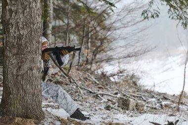 Beyaz kış kamuflajı giyen asker, ağaçların arkasına çömelmiş, sessiz kozalaklı bir ormanda kar taneleri arasından bir tüfeği nişan alıyor. Görüntü gizlilik, hazırlık ve soğuk hava askeri eğitimini aktarıyor