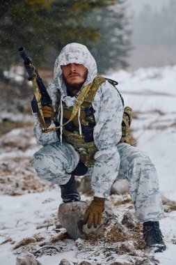 Beyaz kış kamuflajı ve taktiksel yelek giyen silahlı asker, karlı bir ormanda, elinde bir tüfekle bir kayanın üzerine çömelmiş, hazırlık, hayatta kalma ve soğuk hava askeri operasyonlarını sürdürüyor..