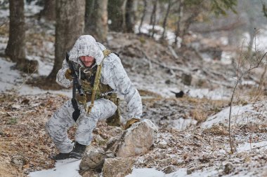 Beyaz kışlık kamuflaj ve taktik teçhizat giyen silahlı bir asker, karlı bir ormanda devriye gezerken bir kayanın arkasına çömelir, soğuk hava askeri operasyonu sırasında gözlem ve hareketlere odaklanır..