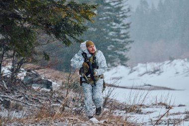 Beyaz kış kamuflajı giyen bir asker, kar ormanlarının kıyısında devriye geziyor, hafif kar yağışı ve bulutlu hava koşullarında keşif yaparken bir tüfek ve taktik teçhizat taşıyor..