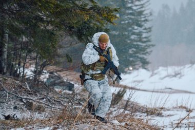 Beyaz kış kamuflajlı bir asker, soğuk hava devriyesi ya da eğitim sırasında elinde tüfekle, karlı bir orman kenarında ihtiyatlı bir şekilde hareket eder..