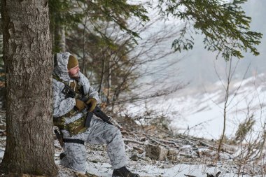 Kış kamuflajı ve taktik teçhizatı giyen asker, karlı bir orman devriyesi sırasında elinde tüfekle bir ağaca doğru diz çökerek ihtiyat, hayatta kalma, hazırlık ve soğuk arazide sessiz gözlem yapıyor..