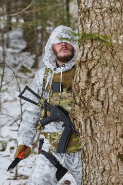 Beyaz kış kıyafetleri ve taktik yelek giyen kamufle olmuş bir asker ağaçların arkasından, katlanmış bir tüfekle ve sessiz, karlı bir ormanda hayatta kalma bıçağıyla silahlanmış..