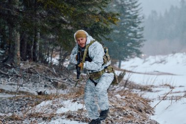 Beyaz kış kamuflajlı ve taktik teçhizatlı bir asker, karlı bir ormanda tüfekle ihtiyatlı bir şekilde ilerler. Düşen kar ve soluk renkler soğuk, ihtiyatlı, hazırlıklılık ve açık hava ordusunu taşır.