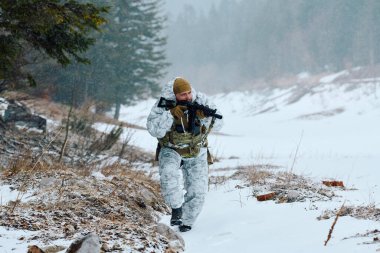Beyaz kışlık kamuflaj ve taktik teçhizat takan bir asker, soğuk hava tatbikatı sırasında kar altında ihtiyatlı bir şekilde ilerleyen bir tüfekle karlı bir orman kenarında devriye geziyor..