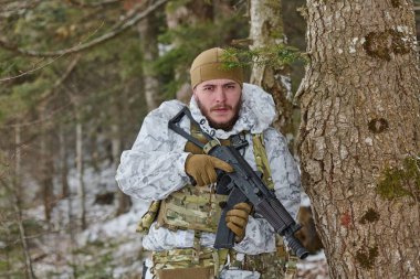 Kış kamuflajlı ve taktik teçhizatlı bir asker, karlı bir ormanda bir ağacın arkasında saklanırken tüfeğini tutuyor. Etrafı odaklanmış ve tetikte bir ifadeyle tarıyor..