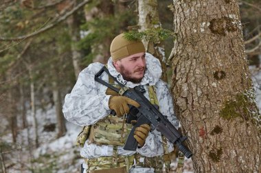 Beyaz kış kamuflajı ve taktik yeleği giymiş bir asker, karlı bir ormanda bir ağacın arkasında saklanırken tüfeğini tutuyor. Hazırlığını, ihtiyatını ve soğuk hava askeri operasyonlarını sürdürüyor..
