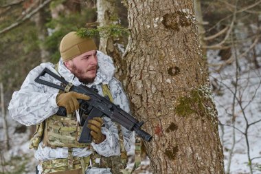 Beyaz kış kamuflajı ve taktik yelek giyen silahlı asker, karlı bir ormanı tararken bir ağacın arkasına saklanıyor. Gizlilik, hazırlık ve soğuk hava askeri devriye operasyonlarını gösteriyor..