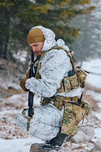 Kış kamuflajlı ve taktik teçhizatlı bir asker, ormanlık alanda soğuk hava tatbikatı sırasında elinde tüfekle karla kaplı zemine diz çöker..
