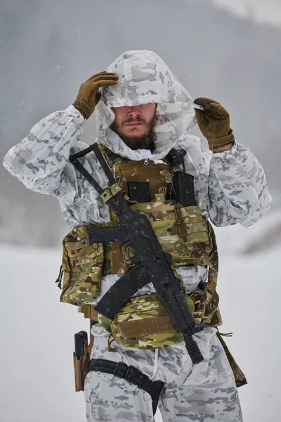 Beyaz kış kamuflajı giyen bir asker, karlı, dağlık bir eğitimde veya devriyede soğuk hava operasyonları sırasında taktik yelek, tüfek ve eldiven ile donatılmış kar yağışı altında başlığını düzeltir.