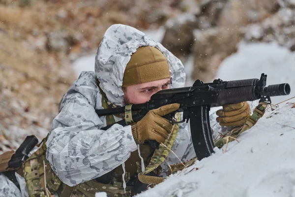 Beyaz kış kamuflajı giyen bir asker soğuk hava askeri tatbikatı sırasında tüfekle kara yatar. Odaklanmış ifade, taktik teçhizat ve düşen kar yoğunluğu iletir