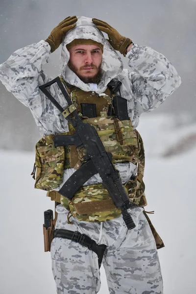 Beyaz kış kamuflajı ve taktik yelek giyen sakallı bir asker, karlı bir eğitim ortamında tüfeğini taşırken kaputunu kavrarken, dayanıklılık, hazırlık ve soğuk hava operasyonlarını iletiyor.