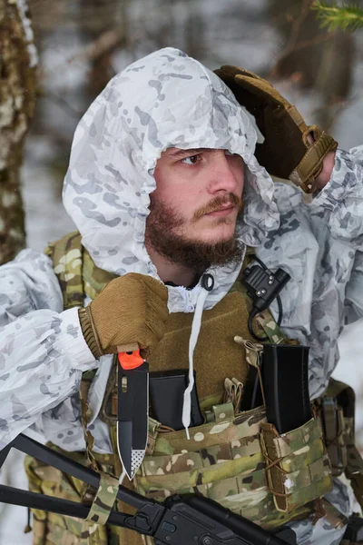 Beyaz kışlık kamuflaj ve taktik yelek giyen sakallı asker, karlı bir ormanda bir tüfeğin yanında hayatta kalma bıçağını tutarken kapüşonunu ayarlıyor. Hazırlık, gizlilik ve soğuk hava koşullarını iletiyor.
