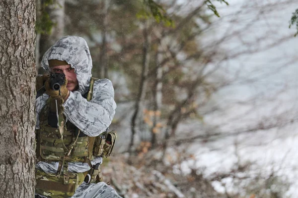 Beyaz kış kamuflajlı ve taktik teçhizatlı bir asker, karlı bir ormandaki bir ağacın arkasından, kar taneleri altında gizlilik, odaklanma ve soğuk hava askeri eğitimini gösteren bir tüfeği hedefliyor..