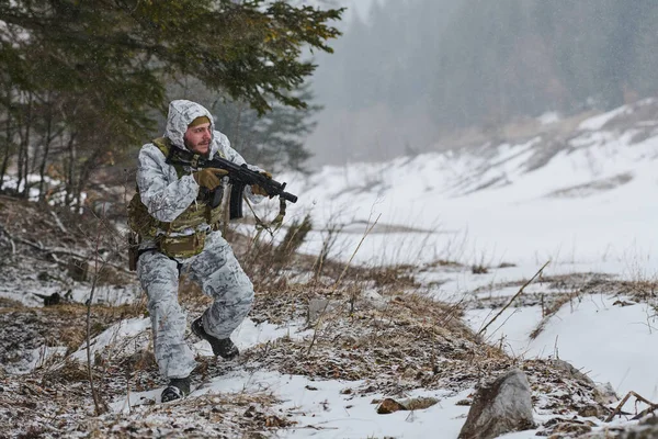Beyaz kışlık kamuflaj ve taktik teçhizat kuşanmış silahlı bir asker, karlı bir orman sınırında dikkatli bir şekilde hareket ederek, engebeli ve ücra bir yerdeki soğuk hava askeri eğitim devriyesi sırasında tüfeğini doğrultuyor.