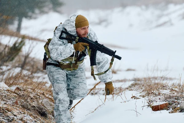 Beyaz kışlık kamuflaj ve taktik teçhizat takan bir asker karlı dağlık arazide devriye geziyor, elinde tüfekle serin bir havada sürüklenirken ve seyrek bir bitki örtüsü altında geziniyor.