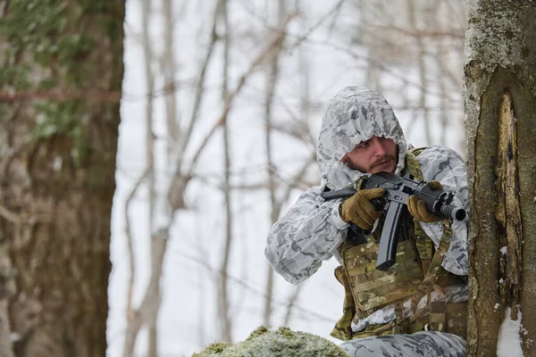 Beyaz kış kamuflajlı asker, karlı bir orman devriyesi sırasında elinde tüfekle bir ağacın etrafını kolaçan ederek taktik gizleme, soğuk hava eğitimi, hazır olma ve orman çatışmasında gizlenmeyi gösteriyor.