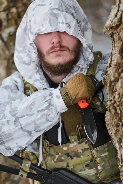 Beyaz kışlık kapüşon ve taktik yelek giyen kamufle asker, karlı bir ormanda bir ağacın arkasında saklanırken bir hayatta kalma bıçağını kavrayıp gizlilik, hazırlık ve soğuk hava askeri operasyonlarını sürdürüyor..