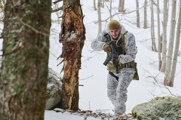 Kış kamuflajlı ve taktik teçhizatlı bir asker, elinde bir saldırı tüfeği tutarken karlı bir ormanda hareket ediyor. Odaklanma, hazır olma ve soğuk hava askeri devriye eğitimi veriyor.