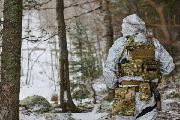 Beyaz kış kamuflajlı bir asker ormandaki soğuk hava operasyonları sırasında gizlenmek ve keşif yapmak için taktik yelek, telsiz ve teçhizat taşıyan karlı bir orman yolunda devriye geziyor.