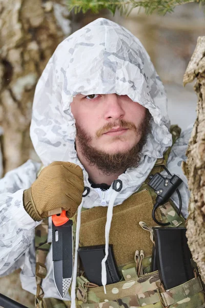 Beyaz kar maskesi takmış sakallı bir asker ve bir taktik plaka taşıyıcısı bir kış ormanında ağaçlara karşı dikilirken bir savaş bıçağı tutuyor..