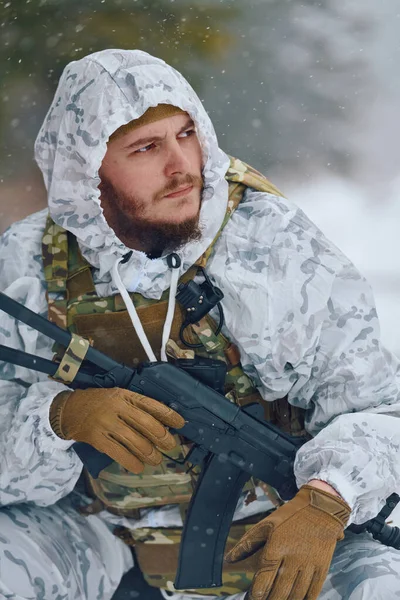 Beyaz kış kamuflajlı silahlı bir asker karlı bir ormanda çömelmiş, bir tüfek tutuyor ve taktik yelek ve eldiven giyiyor. Huysuz, ihtiyatlı bir sahne, hayatta kalma, hazırlık ve soğuk havayı anlatıyor.