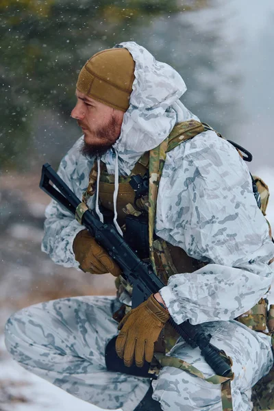 Beyaz kış kamuflajlı ve taktik teçhizatlı bir asker kar yağarken çömelir, soğuk hava devriyesi sırasında karlı bir ormanı incelerken tüfeği kavrar ya da tatbikat yapar..