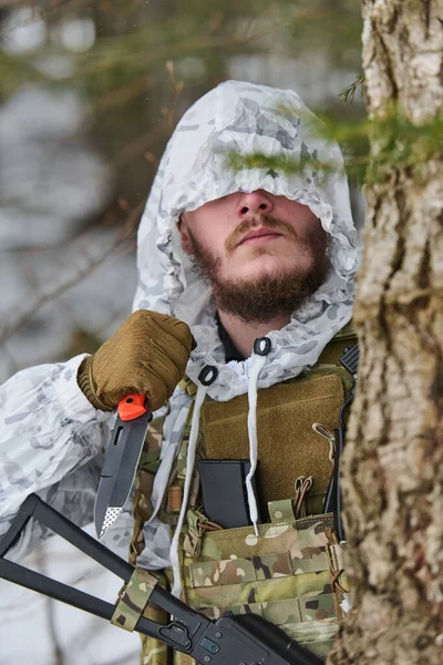 Beyaz kış kamuflajı ve taktik yelek giymiş sakallı bir asker, karlı bir ormanda bir ağacın arkasında gizlenirken tüfeğin yanında bir savaş bıçağını kavrıyor. Gizlilik, hazırlık ve hayatta kalma gerginliğini çağrıştırıyor..