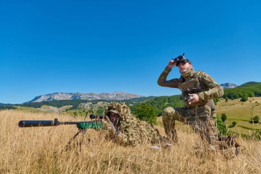 Kamuflajlı iki asker, gulyabani kıyafetli bir keskin nişancı ve FPV gözlüklü bir İHA operatörü kayan tepeleri ve açık mavi gökyüzü olan kuru bir çim tarlasında keşif ve eğitim yapıyorlar..
