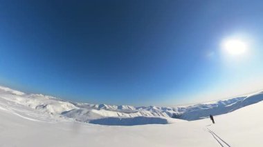 Kıdemli bir freeride kayakçısı, heyecan verici bir pist dışı helikopter pisti turu sırasında taze ve derin toz karda zarif çizgiler çiziyor. Görkemli karlı dağ manzarası berrak mavi bir gökyüzünün altında açılır.
