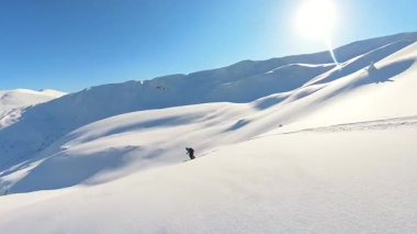 Taze, derin toz karla kaplı el değmemiş bir dağda kayak yapmanın heyecanını yaşayın. Solo bir kayakçı parlak mavi gökyüzünün altında zarif dönüşler yapar, özgürlüğünü gözler önüne serer.