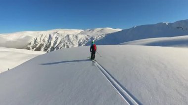 Kıdemli bir kayakçı, uçsuz bucaksız bir dağ yamacında zarif bir şekilde el değmemiş, derin toz kar yağışının tadını çıkarıyor. Parlak kış güneşi karlı havayı aydınlatıyor.