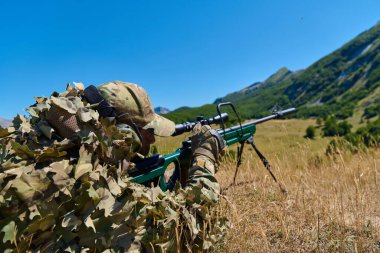 Kamuflajlı keskin nişancı asker, dağlık bir vadide iki ayaklı dürbünlü dürbünlü dürbünlü tüfeği hedef alarak kuru çimenlerde yatar. Açık hava askeri eğitim sahnesi. Gizlilik, hassasiyet, uzun menzilli.