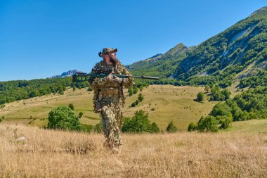 Kamuflajlı keskin nişancı asker elinde hassas bir tüfekle dağlık bir çayırda yürüyor. Üzerinde ghillie tarzı kamuflaj ve taktik teçhizat var. Parlak yaz manzarası hazırlığı ve gizliliği akla getiriyor.