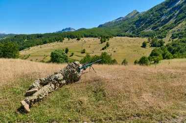 Kamuflajlı keskin nişancı asker, güneşli bir dağ vadisinde hassas bir tüfeği hedefliyor. Eğitim tatbikatı sırasında çimenli çayırlarda taktik gizleme ve uzun menzilli keşif.