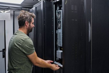 Bir IT teknisyeni veri merkezi rafı içinde sunucu donanımı inceler ve korur, sistem güvenilirliğini, soğutma verimliliğini ve ağını sağlamak için bileşenleri yönetir ve rutin bakım yapar