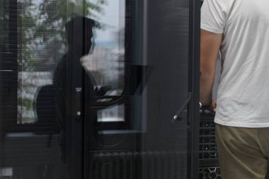 Bir bilişim teknisyeni güvenli bir veri merkezindeki sunucu rafındaki donanımı düzeltirken, bir meslektaşı da cam kapıya yansıyarak takım çalışması, bakım, altyapı ve ağ operasyonlarını aktarıyor..