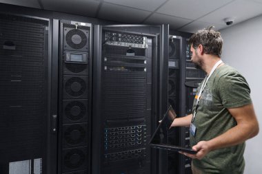 Bir bilişim teknisyeni, yüksek teknolojili bir ortamda bakım, konfigürasyon ve ağ sorunları yaratmak için bir dizüstü bilgisayar kullanarak güvenli bir veri merkezinde sunucu raflarını inceler..